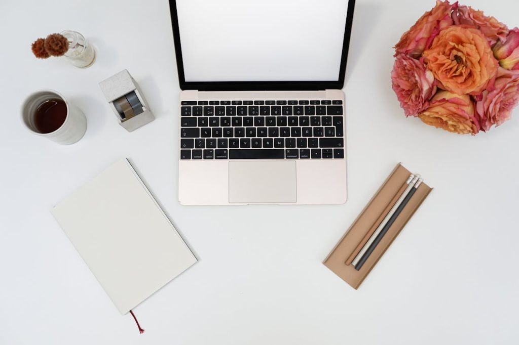 Top view of modern laptop with blank screen arranged on table with stationery and notebook near bunch of fresh flowers and cup of aromatic coffee